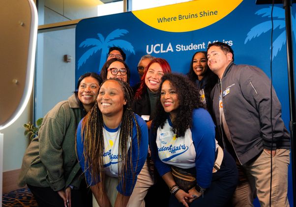 A group of nine people pose for a shot in front of a blue-and-gold sign that reads “Where Bruins Shine” and “UCLA Student Affairs”