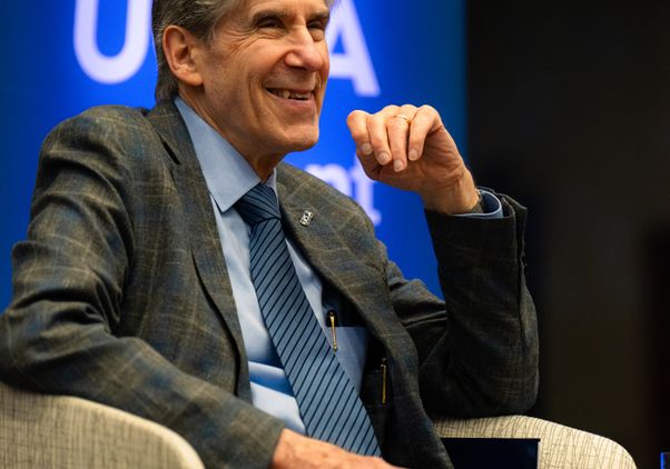 Julio Frenk, in a gray suit, smiles as he sits in a cream-colored chair