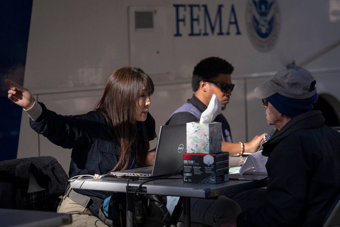 At right, a man in sunglasses and a cap listens as a woman in front of a laptop computer gestures with her hand pointing to the right.
