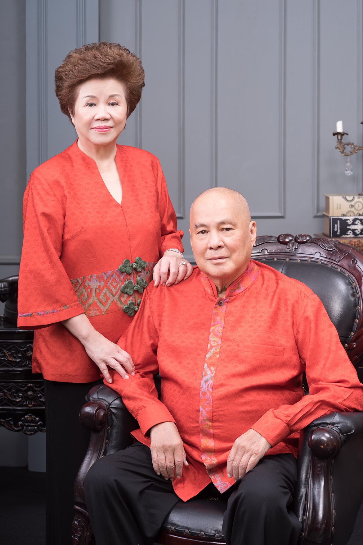 Formal portrait in coordinated red attire: Lelarati Lukman, left, stands with one hand resting on Karmaka Surjaudaja shoulder, while the Surjaudaja sits in a dark leather armchair, both facing the camera