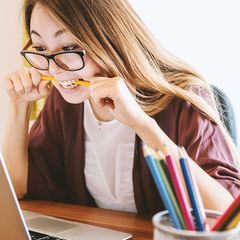 A young woman with glasses chews on a pencil while looking at a laptop, with colored pencils on the desk beside her.