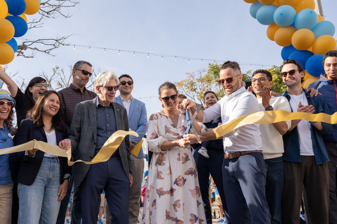 UCLA Chancellor Julio Frenk and Katy Young Yaroslavsky smile in a group as Westwood Village Improvement Association’s Michael Russell uses large scissors to cut horizontal yellow ribbon, in front of blue-and-yellow balloon arch
