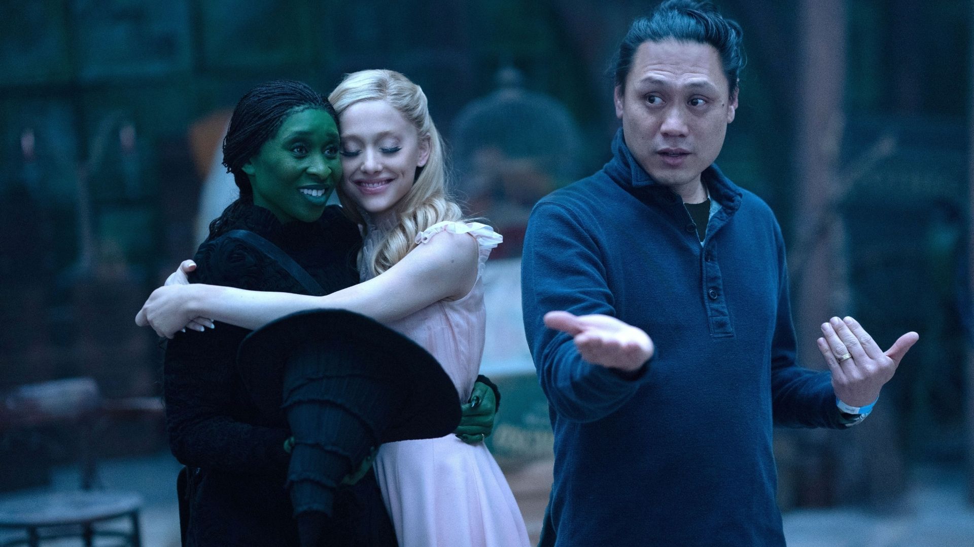 Director Jon M. Chu gestures while giving instructions on the set of "Wicked" as actresses Cynthia Erivo and Ariana Grande embrace in the background.