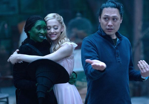 Director Jon M. Chu gestures while giving instructions on the set of "Wicked" as actresses Cynthia Erivo and Ariana Grande embrace in the background.