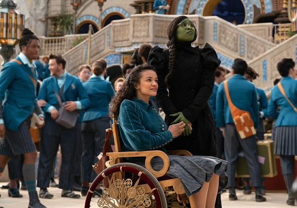 Actresses Marissa Bode, seated in an ornate wooden wheelchair, and Cynthia Erivo in green makeup stand together in a crowded school courtyard scene from "Wicked."