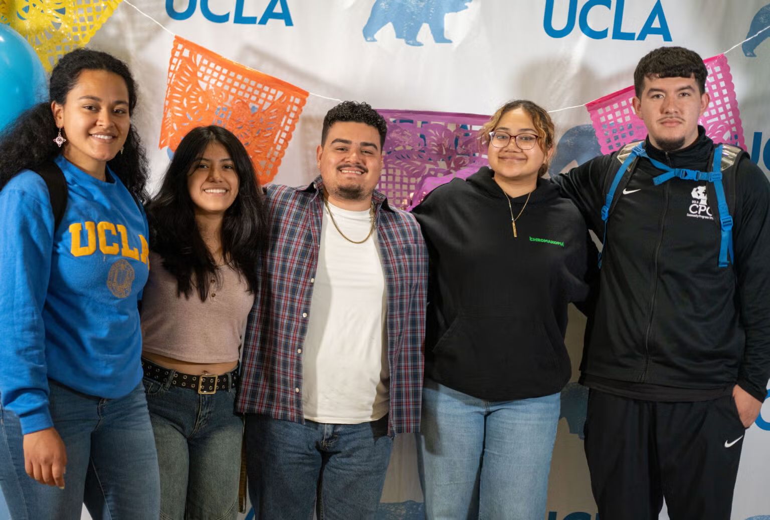Four UCLA students smile and pose together in front of a UCLA banner decorated with colorful papel picado at the opening of UCLA's Latinx Success Center on Cesar Chavez Day. One student wears a UCLA sweatshirt.