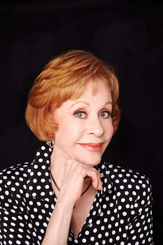 Headshot of Carol Burnett with short auburn hair, wearing a black and white polka-dot blouse, her hand resting under her chin, against a dark background.