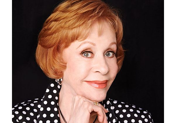 Headshot of Carol Burnett with short auburn hair, wearing a black and white polka-dot blouse, her hand resting under her chin, against a dark background.