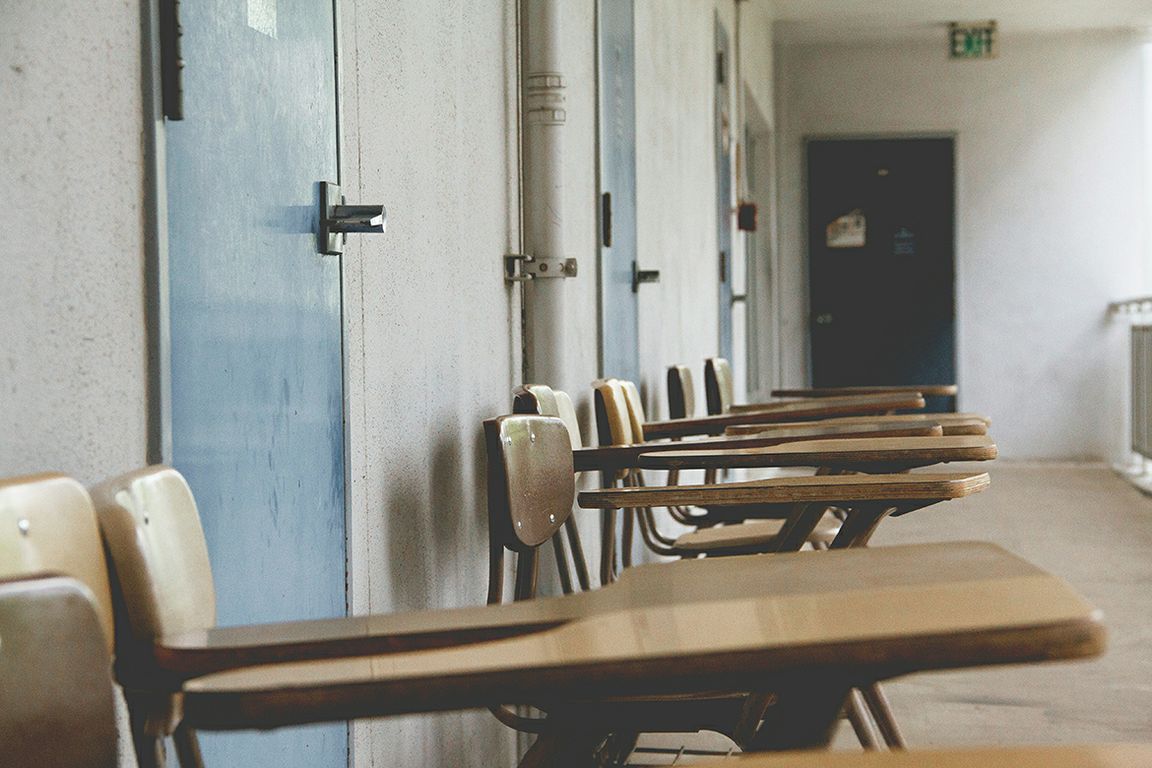 A row of empty wooden student desk-chairs lines a school corridor next to blue-painted lockers, with an exit sign and a door visible in the background.
