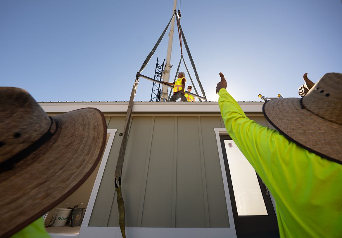 A low-angle view looking up at construction workers in safety vests on the roof of a prefab home being lowered by crane under a clear blue sky. A worker in the foreground wearing a neon green shirt and straw hat gives a thumbs up.