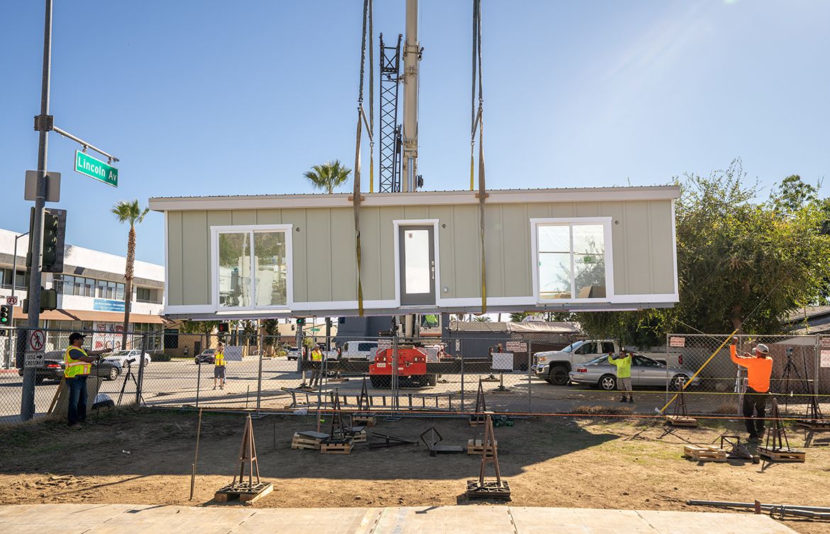 A crane lowers a complete prefab home module onto a fenced construction site at the corner of Lincoln Avenue in Altadena, California. Several workers in safety vests guide the placement while palm trees and street signs are visible in the background. 