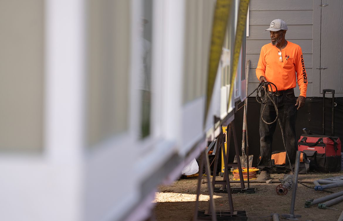 A construction worker in an orange long-sleeve shirt and white cap holds rigging rope while standing beside a prefab home exterior in Altadena. Construction tools and equipment are visible on the ground around him.
