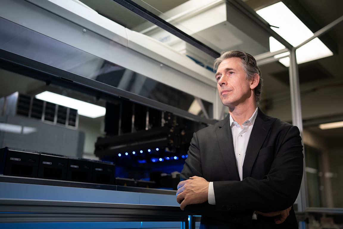 Professor Damoiseaux stands with arms crossed next to the CellXpress.AI automated laboratory instrument, which glows with blue LED lighting, inside a research facility.