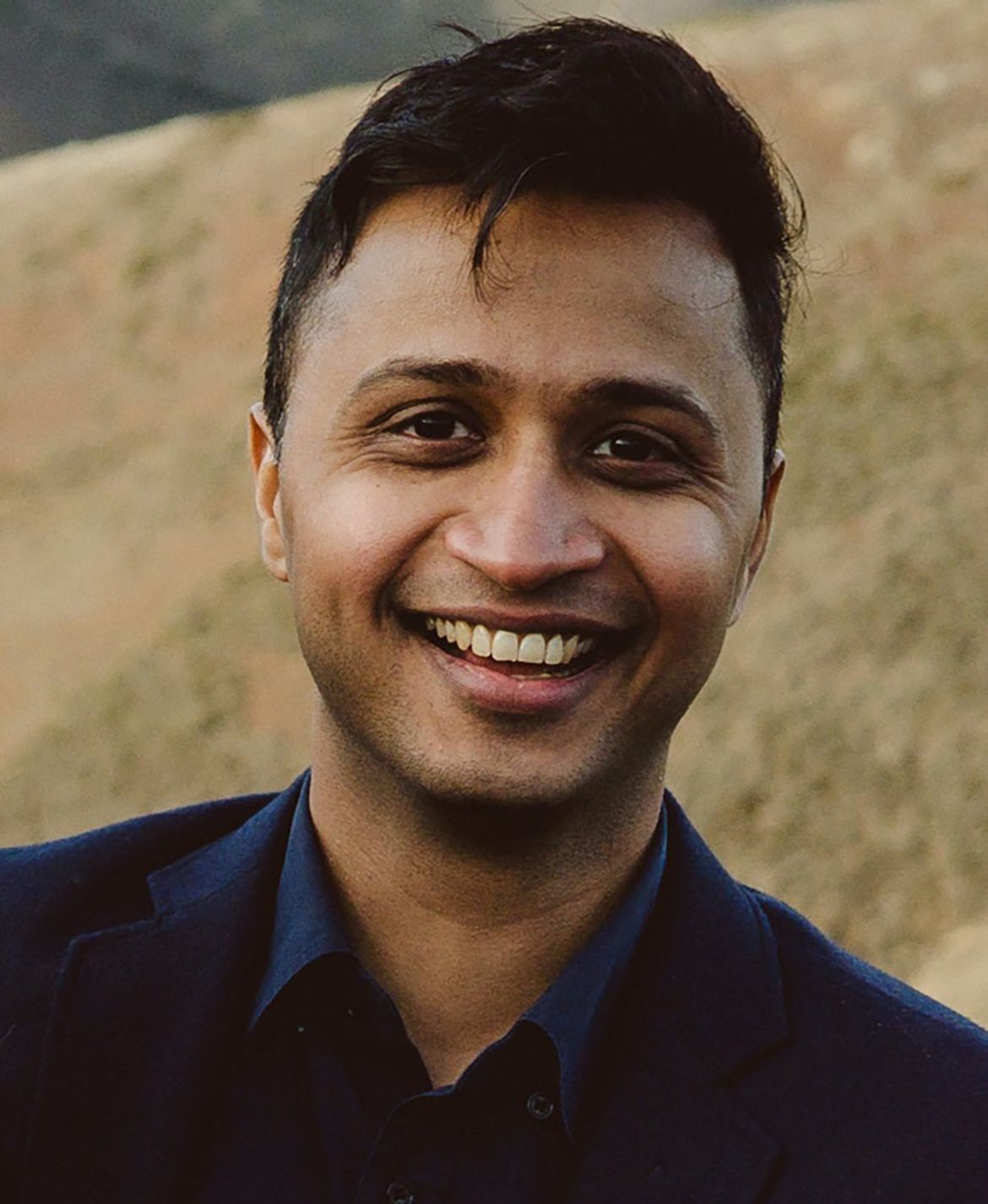 Headshot of Rachit Dubey smiling and wearing a dark navy button-up shirt, with a blurred rocky outdoor background.