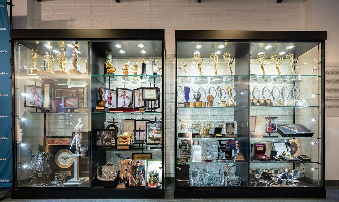 Two illuminated glass display cases filled with Carol Burnett's awards donated to UCLA, including multiple Emmy statuettes on the top shelves, a Grammy, her Kennedy Center Honor, and numerous other trophies, plaques, and memorabilia.