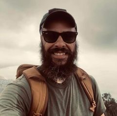 Brandon Hervey takes a selfie outdoors under an overcast sky. He has a full beard, wears dark sunglasses and a baseball cap, and carries a backpack over a gray-green shirt. 