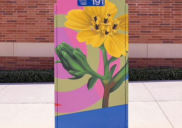  A painted electrical box numbered 191 displays a bold yellow flower with green stem and bud against a pink and olive background with a blue stripe at the bottom, set against a red brick wall on the UCLA campus.