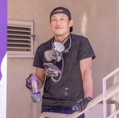 L.A.-based muralist John Park stands outdoors holding a spray paint can, wearing a gray t-shirt, backward baseball cap, and a respirator mask around his neck.