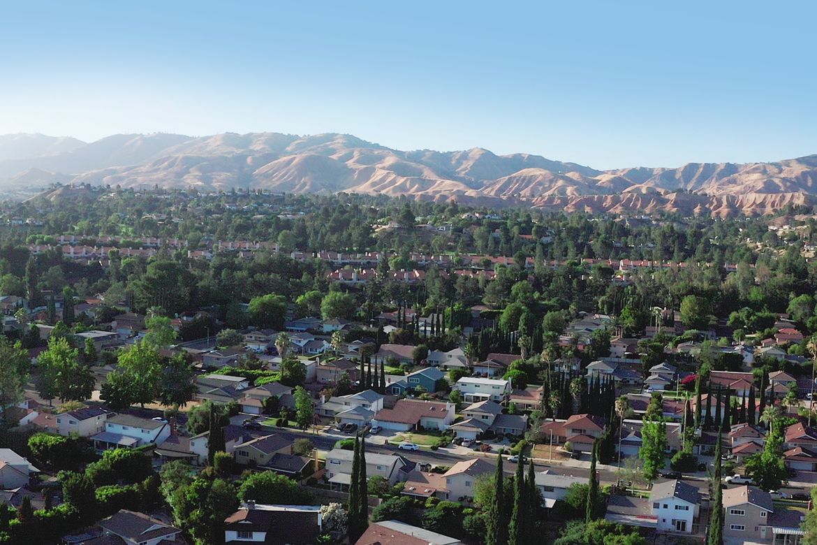 Aerial view of the Aliso Canyon neighborhood showing a dense residential area with single-family homes, tree-lined streets, and green vegetation, set against dry brown hills and a clear blue sky in the background.