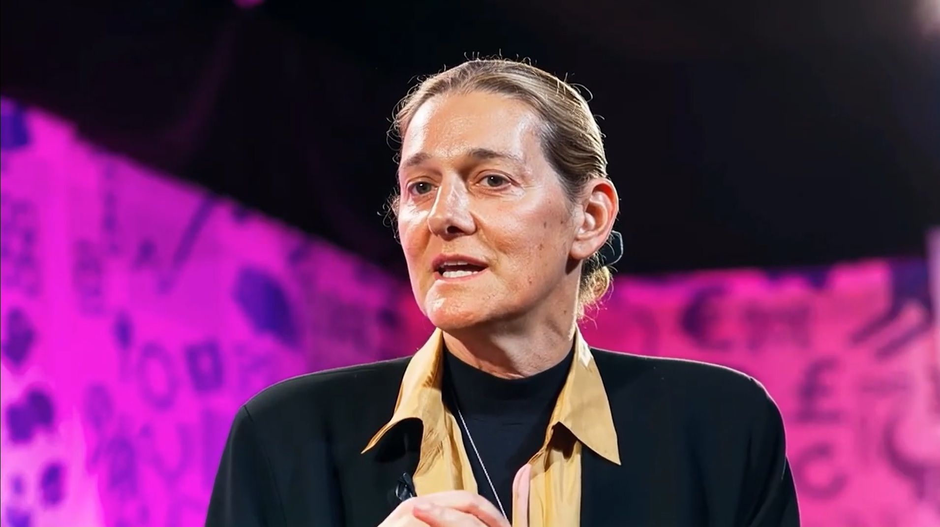Martine Rothblatt speaks at an event. She wears a black top with a gold-collared jacket and a thin necklace with a small pendant.