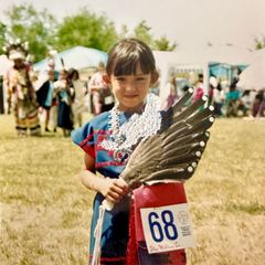 A vintage photograph of a young Tazbah Rose Chavez as a child, dressed in traditional Native American regalia, including a blue dress, white beaded collar, and feathered fan, wearing competition number 68 at an outdoor powwow.