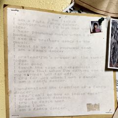 A typewritten first-grade poem by Tazbah Rose Chavez, titled "I Am Tazbah," pinned to a yellow wall alongside small personal photos. The poem describes her identity as a Piute fancy dancer, with lines about powwow music, ceremony, and the joy of dancing. A small school portrait photo is pinned to the upper right corner of the poem.