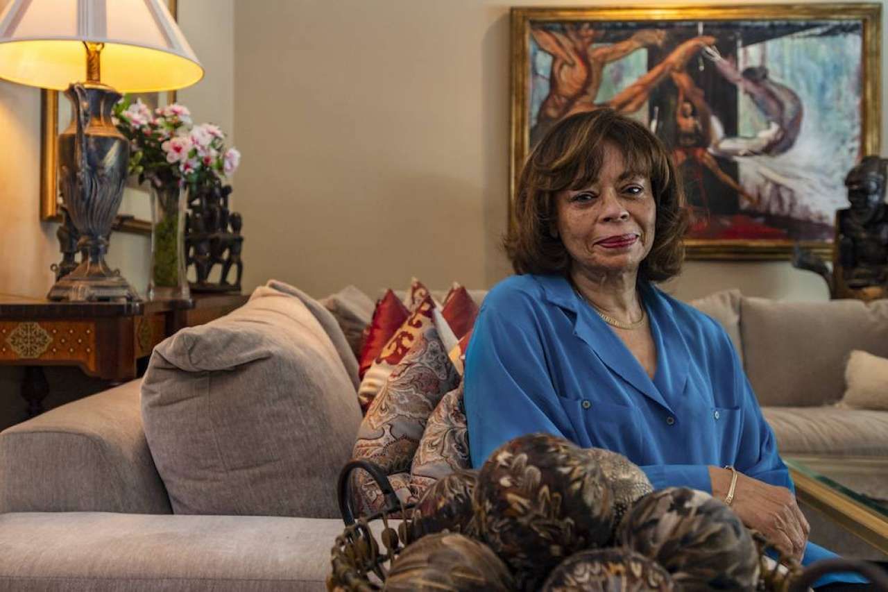 Dr. Gail Wyatt sits on a sofa in an elegantly decorated living room, wearing a blue blouse and gold necklace, with colorful pillows visible in the background.