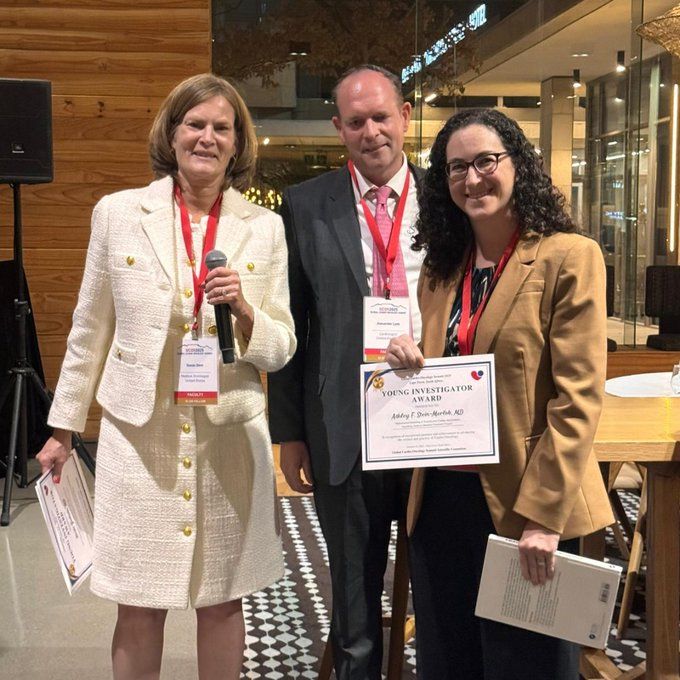 Dr. Ashley Stein-Merlob stands with two colleagues at an awards event, holding a Young Investigator Award certificate, all wearing conference lanyards in an indoor venue