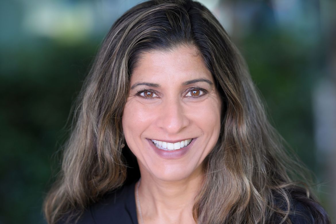 Headshot of Dr. Shafali Jeste smiling, with long brown highlighted hair, against a soft green outdoor background. 