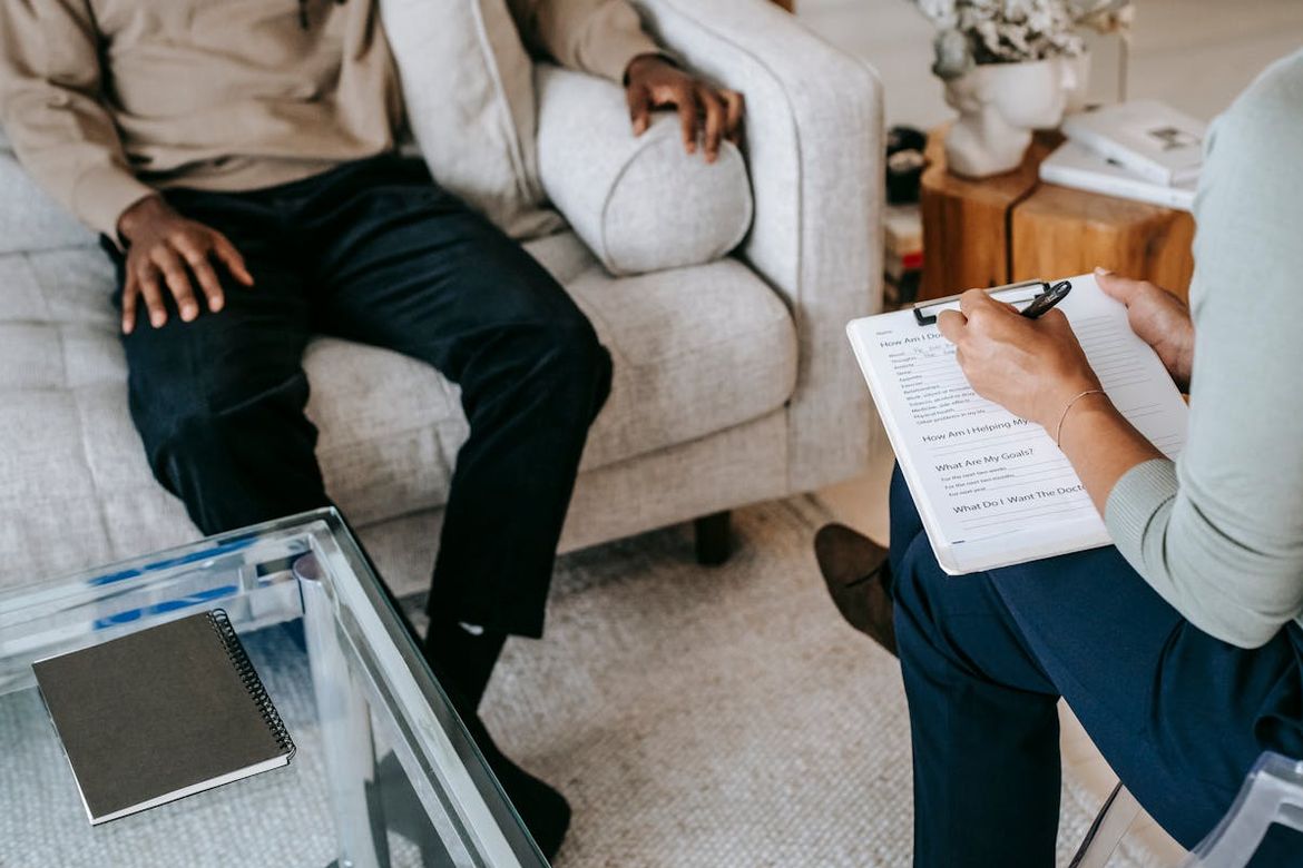 Legs of man seated on beige couch across from female with clipboard in a chair