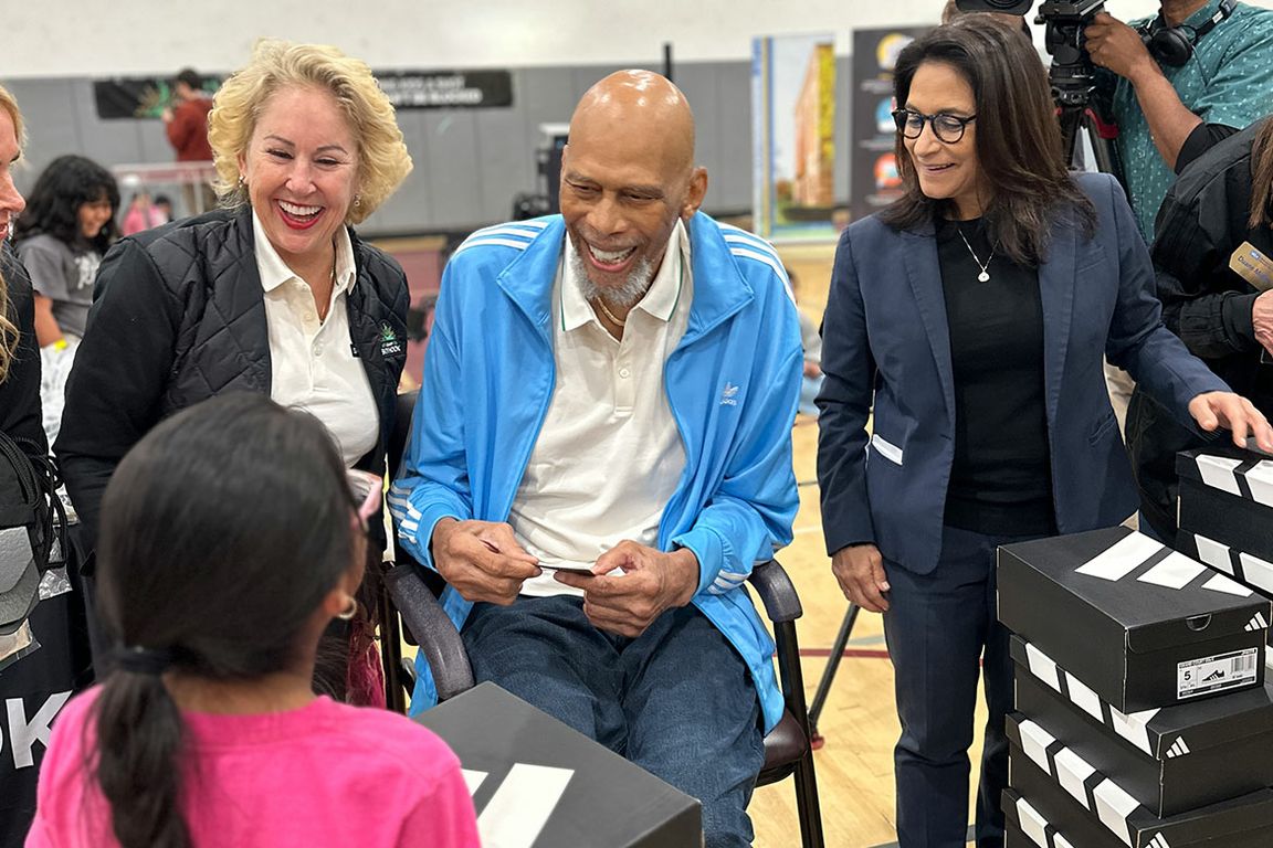 Kareem Abdul-Jabbar, seated and wearing a blue Adidas track jacket, smiles warmly at a young student during a shoe giveaway event in a school gymnasium, flanked by two smiling women, with stacked shoe boxes visible nearby.