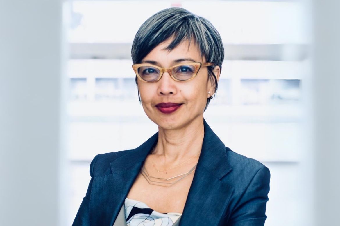 Headshot of Ninez Ponce smiling, wearing cat-eye glasses, a dark blazer, patterned top, and a delicate gold necklace, with a soft bright background.