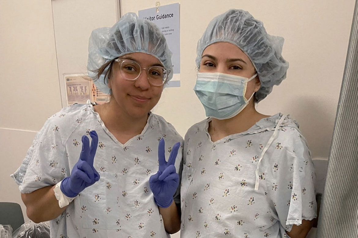 Sisters Rosa (left) and Karina (right) pose together in hospital gowns and surgical caps before their kidney donation procedure at UCLA. Rosa holds up two peace signs with blue-gloved hands, while Karina wears a surgical mask.