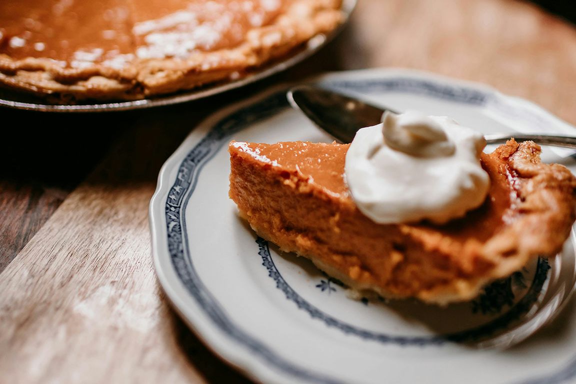 A close-up of a slice of pumpkin pie topped with whipped cream on a blue-and-white decorative plate, with a whole pumpkin pie in a tin visible in the background on a wooden surface