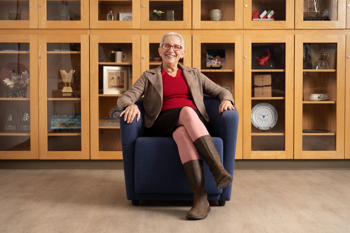 Kathryn Paul smiles while seated in a blue armchair, wearing a red sweater, tan blazer, pink tights, and brown boots, in front of a wall of wood-and-glass display cabinets holding awards and memorabilia.
