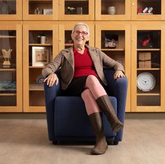 Kathryn Paul sits smiling in a blue armchair in front of wooden display cases filled with awards and mementos.
