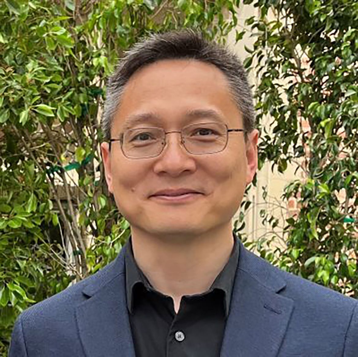 Headshot of Zhongbo Kang smiling, wearing glasses, a black shirt, and a navy blazer, with green foliage in the background.