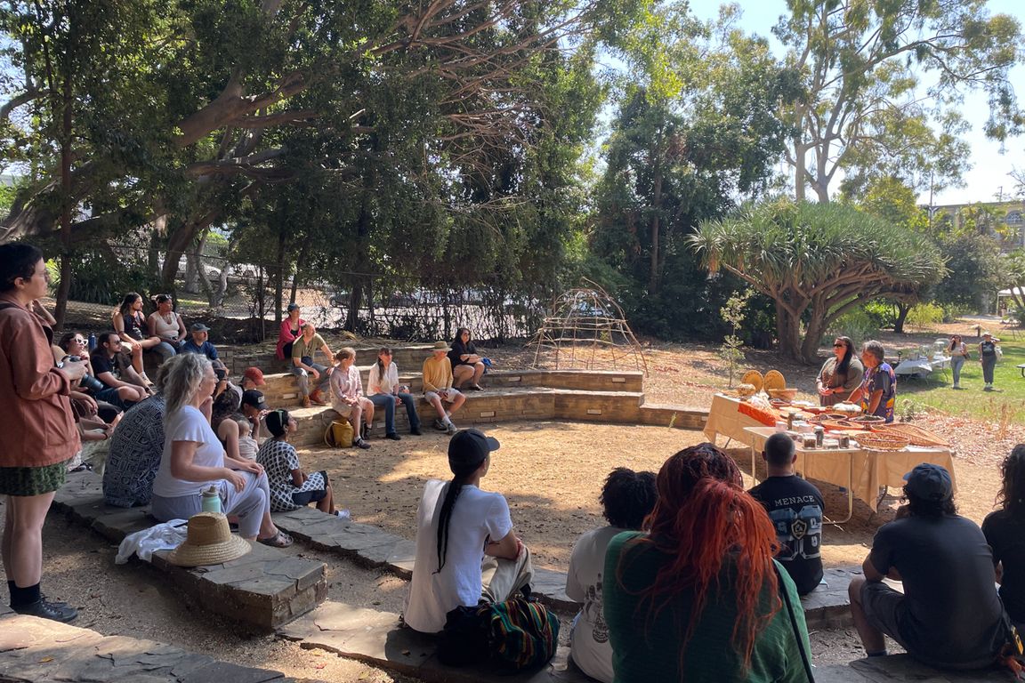 About 20 people sit in a semicircle on stone steps outdoors under trees, watching Theresa Ambo lead a teaching session near a display table at Kuruvungna Springs, West Los Angeles.