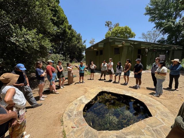 About 15 people stand in a semicircle around a stone-rimmed natural spring pool outdoors, listening to an instructor speak at Kuruvungna Springs, West Los Angeles. Trees and a green building are visible in the background.