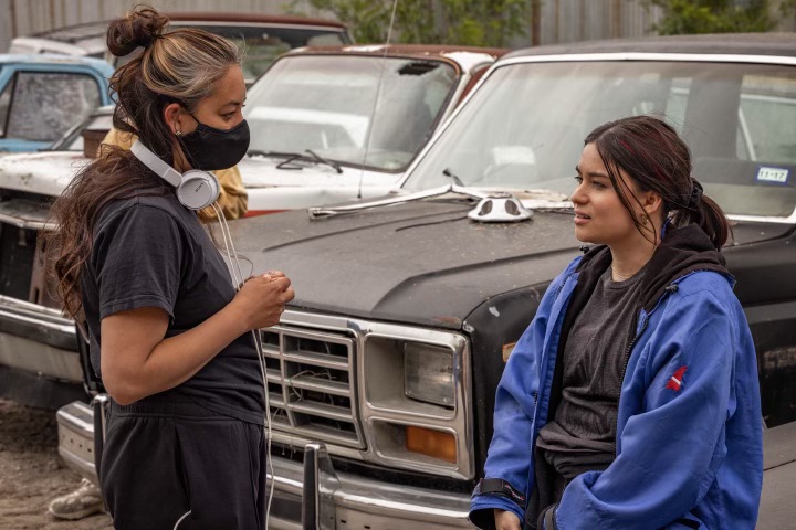Director Tazbah Rose Chavez, wearing a black mask and white headphones, speaks with an actor on the outdoor set of "Reservation Dogs." They stand in front of a weathered black truck.