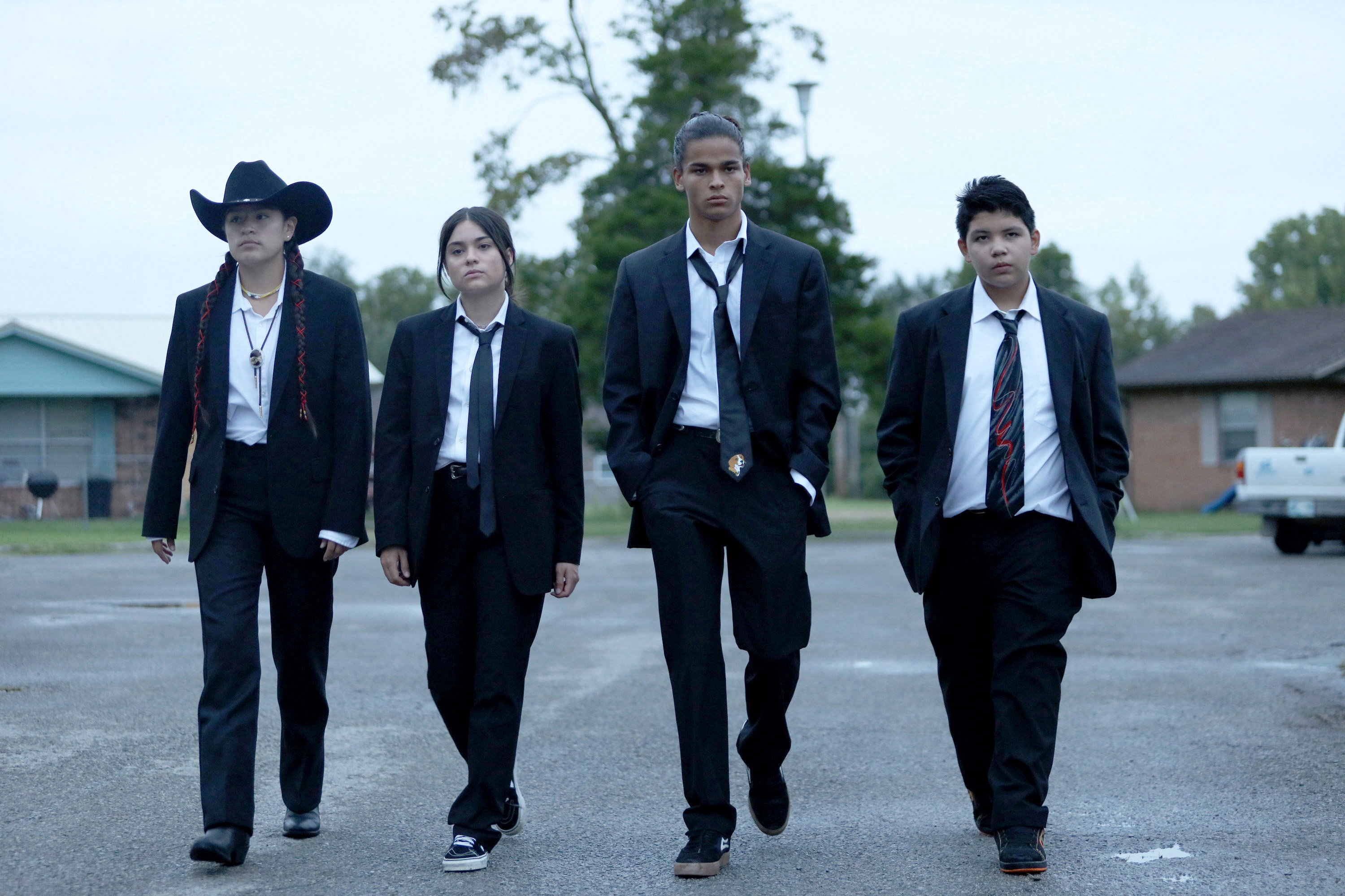 Four young cast members of the FX series "Reservation Dogs" walk confidently side by side in matching black suits, white shirts, and ties. The leftmost person wears a black cowboy hat with long braids; the others wear their hair down or pulled back. They walk through a parking lot with residential buildings and trees in the background.