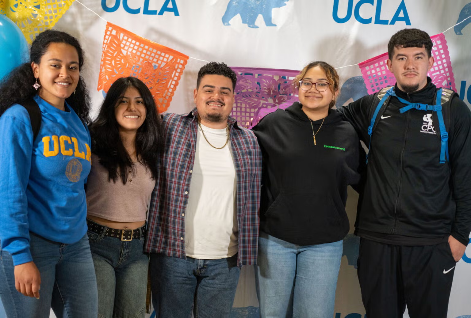 Five students stand in front of a white step-and-repeat with multicolored papel picado hands overhead.