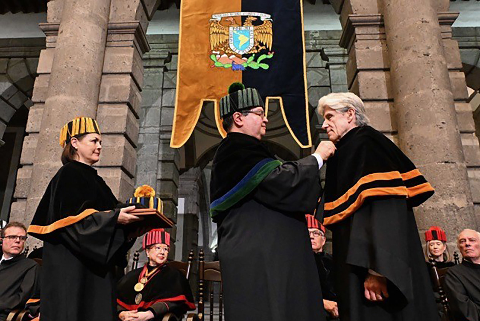 Woman, left, holds diploma covers and hat as man, center, adjusts Julio Frenk's cape
