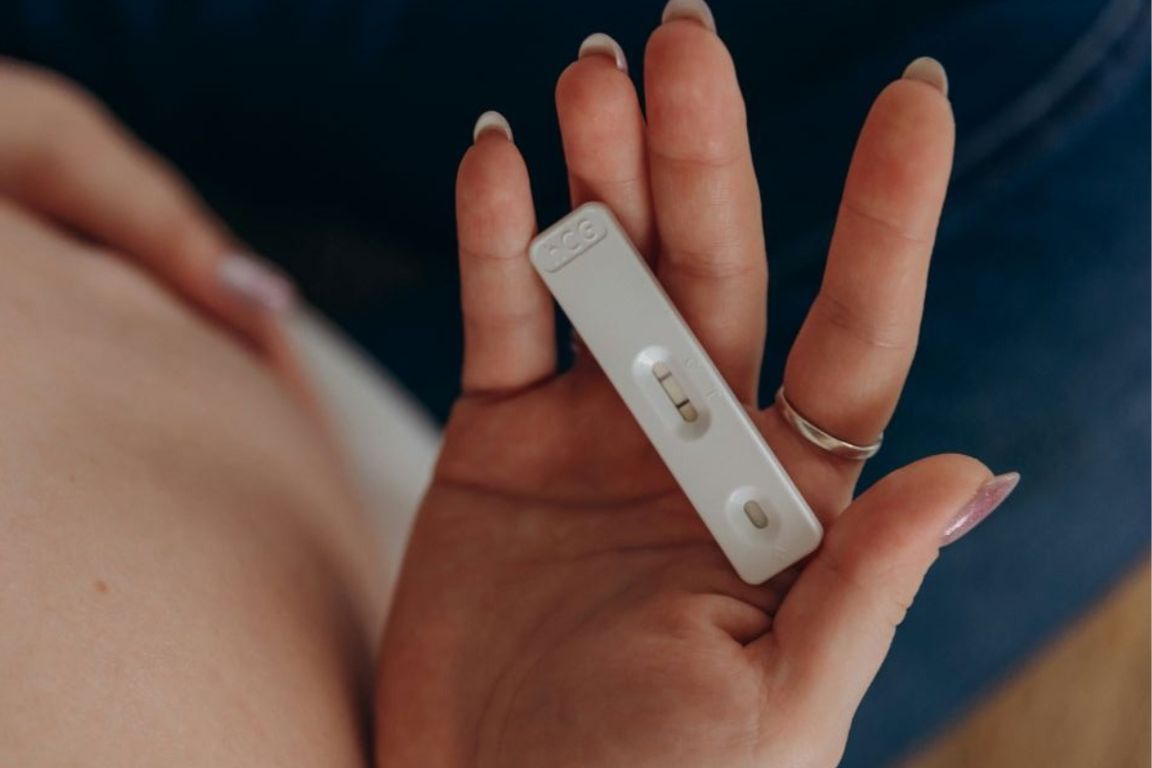 A right hand holds a white pregnancy test with two lines over a bare pregnant belly