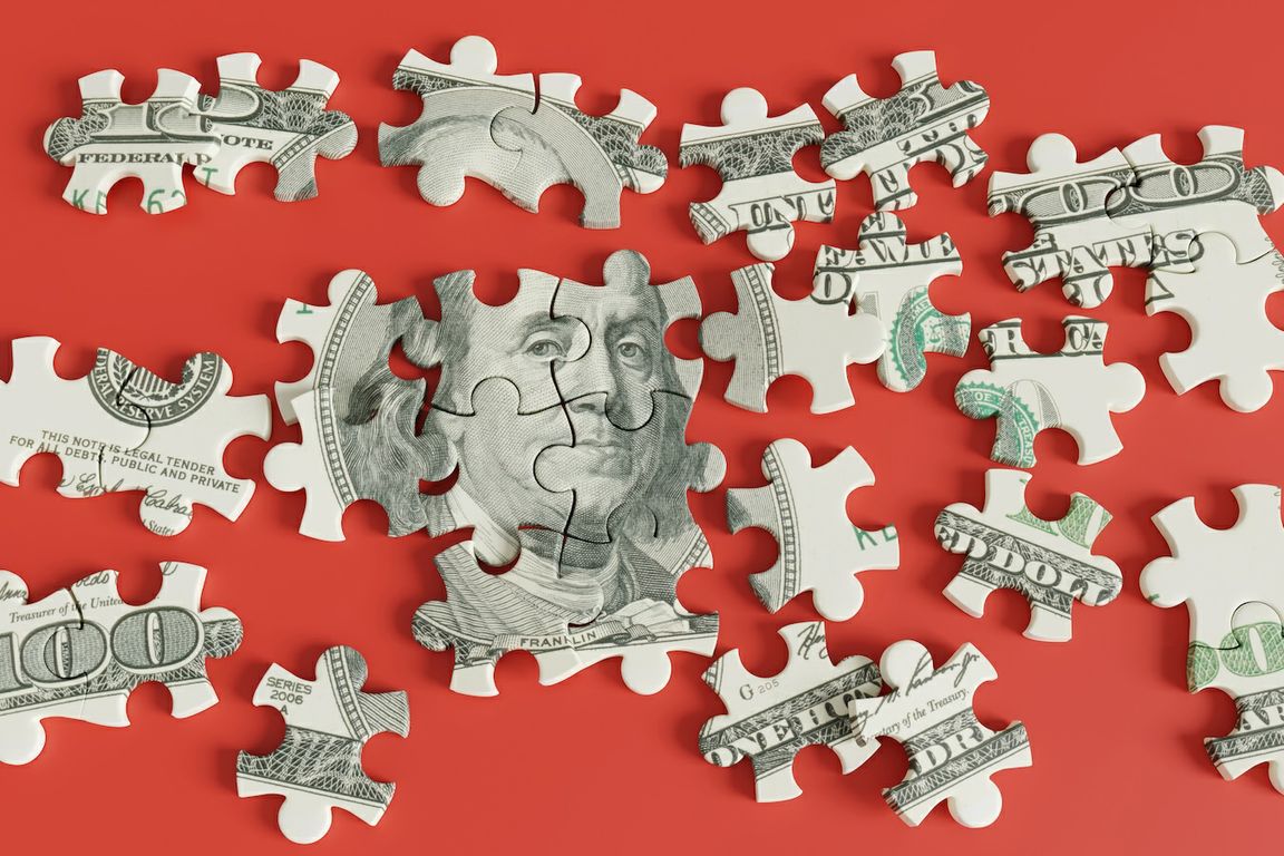A partially assembled puzzle made from a U.S. hundred-dollar bill, with scattered pieces on a red background.