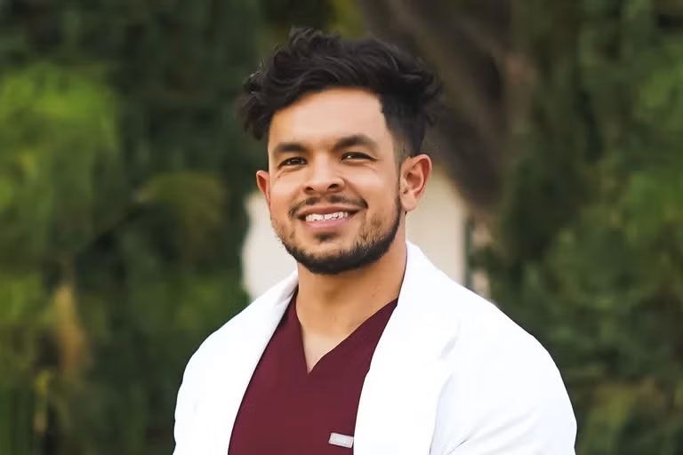 Headshot of Alexander Sanchez smiling outdoors, wearing burgundy scrubs and a white lab coat, with green trees in the background.