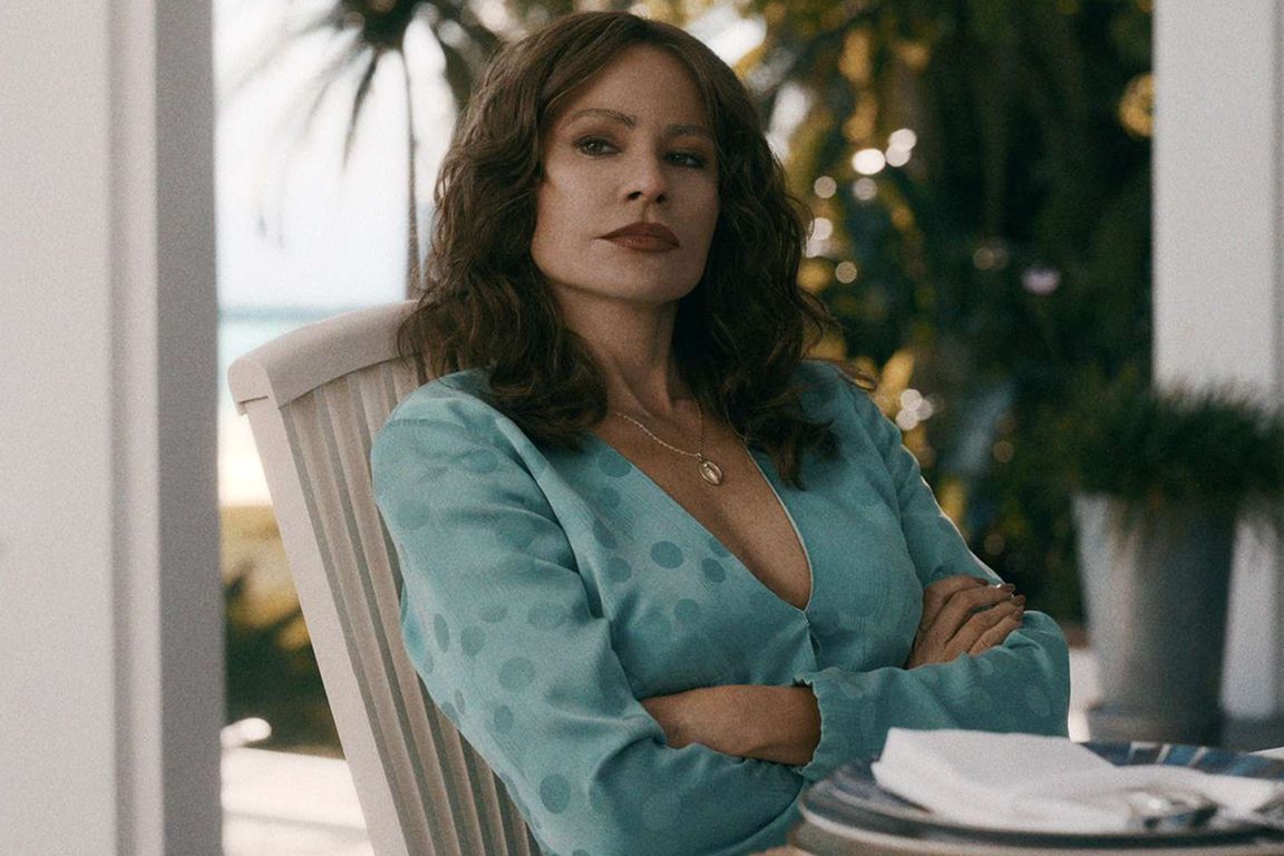 Sofia Vergara as Griselda in Netflix's "Griselda," seated outdoors with arms crossed, wearing a teal polka-dot blouse, with a ocean view in the background.