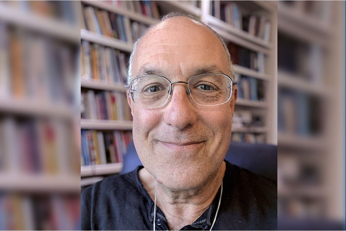 Close-up of Ron Avi Astor, in dark shirt and glasses, sitting in front of a bookshelf