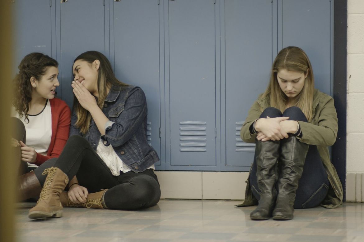 Two teenage girls sit together against school lockers laughing, while a third girl sits apart from them, hugging her knees and looking down, appearing isolated.