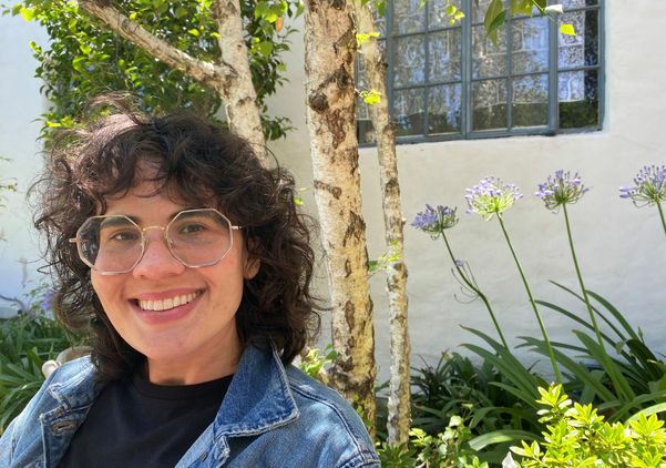 Headshot of Cailey Hall smiling outdoors, wearing glasses and a denim jacket, with trees and purple flowers in the background. 